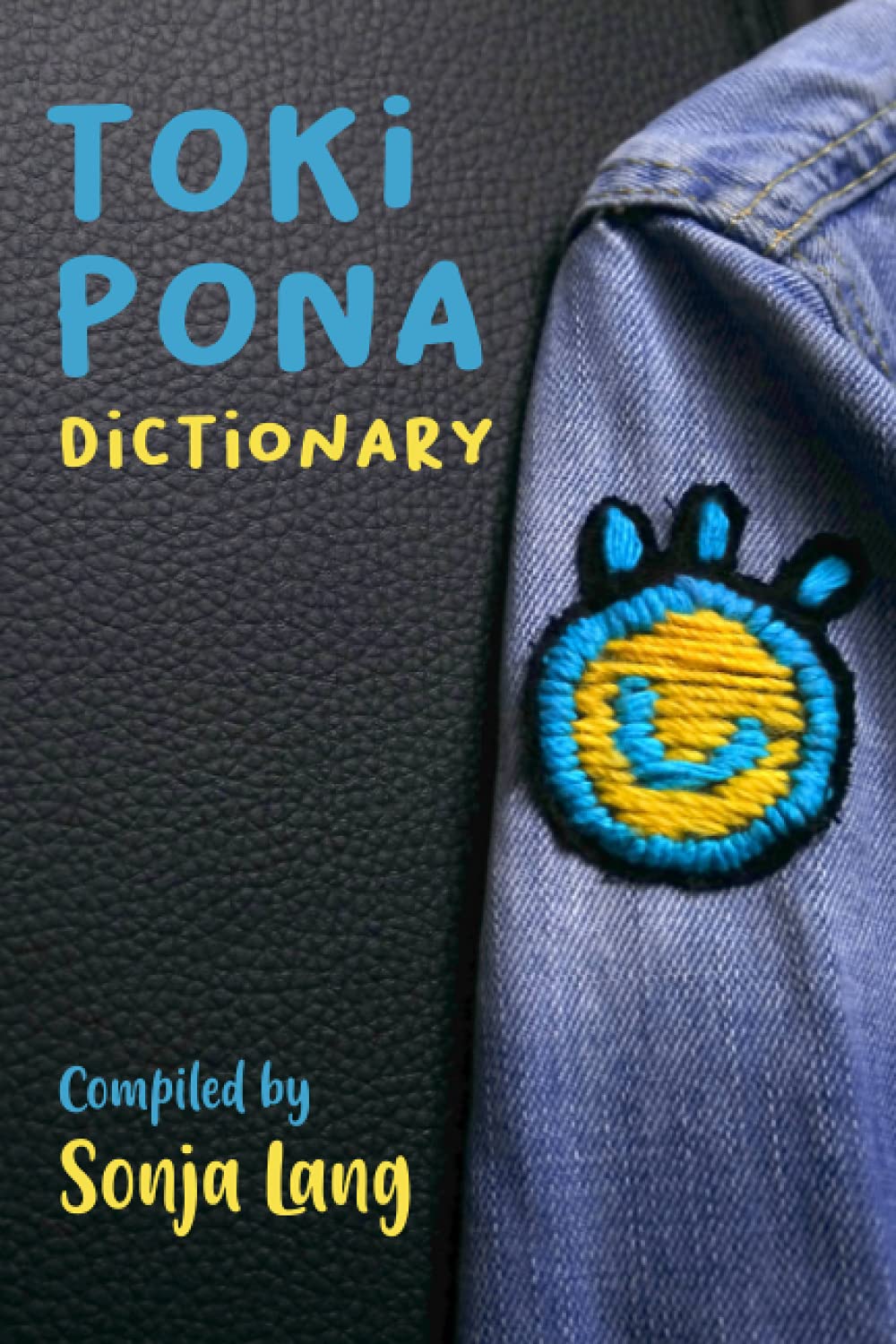 The text 'toki pona' is in blue at the top left. Below it reads 'dictionary' in yellow. To the right of it all is a shirt with the toki pona logo on it.